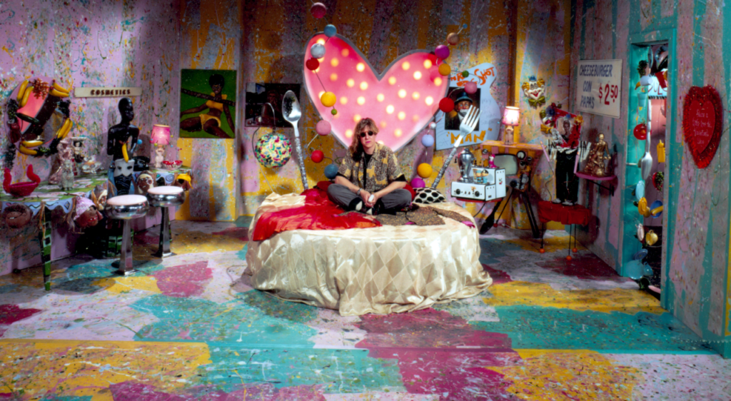 A person sits on a bed with a large heart-shaped light fixture in a colorful, eclectic room filled with various art pieces and decorations.