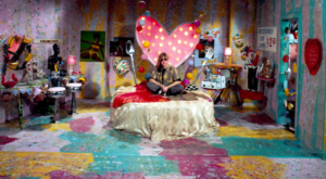 A person sits on a bed with a large heart-shaped light fixture in a colorful, eclectic room filled with various art pieces and decorations.