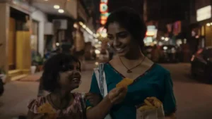 A woman and child smile while walking on a city street at night, holding fried snacks in their hands.