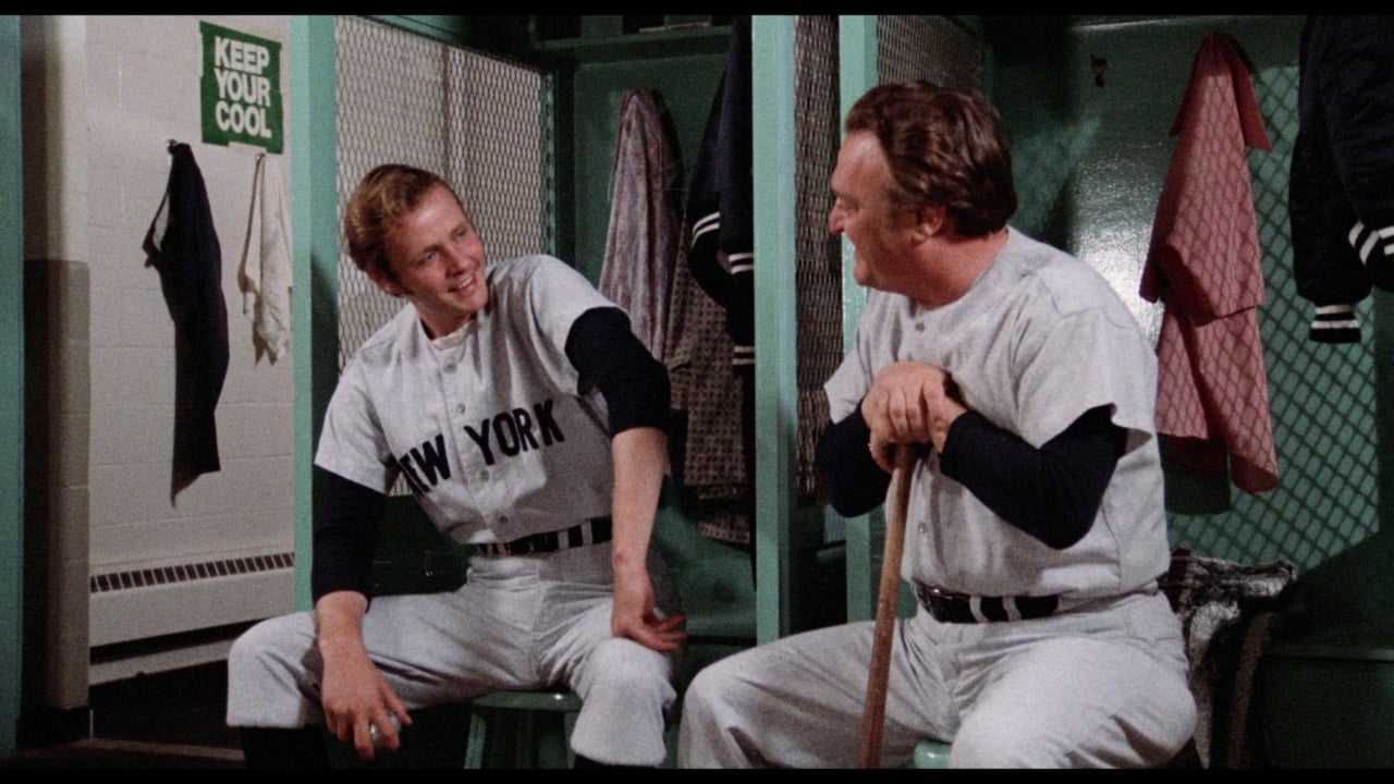 Two baseball players in New York uniforms sit and converse in a locker room. One holds a bat. The walls have hanging jackets and a "Keep Your Cool" sign is visible.
