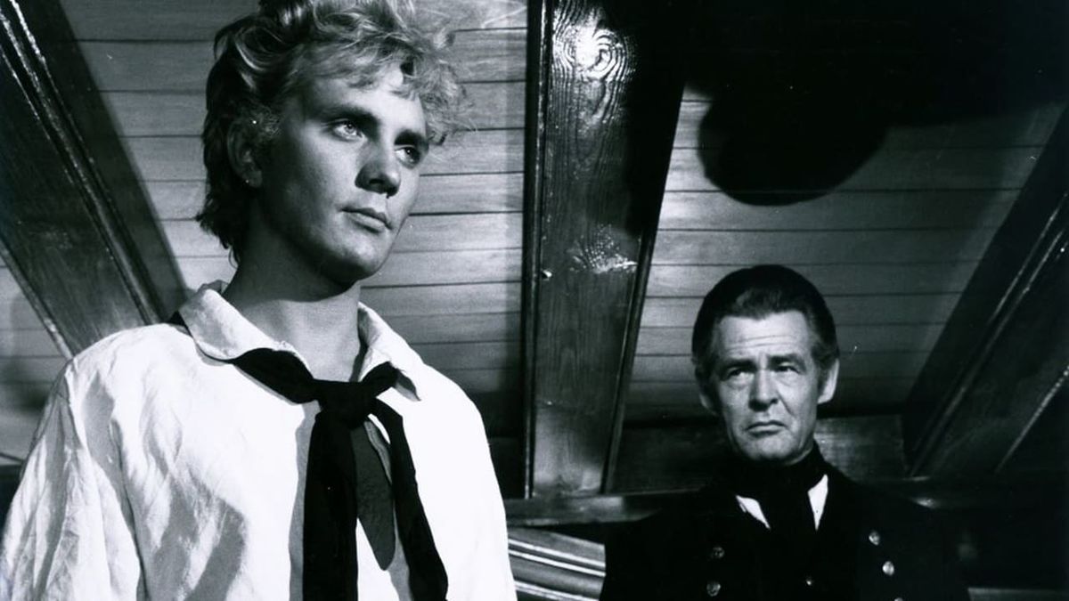 Two men in naval uniforms stand on a ship. The one in front has blond hair and a white shirt, while the man behind wears a dark coat. The setting suggests an older period.