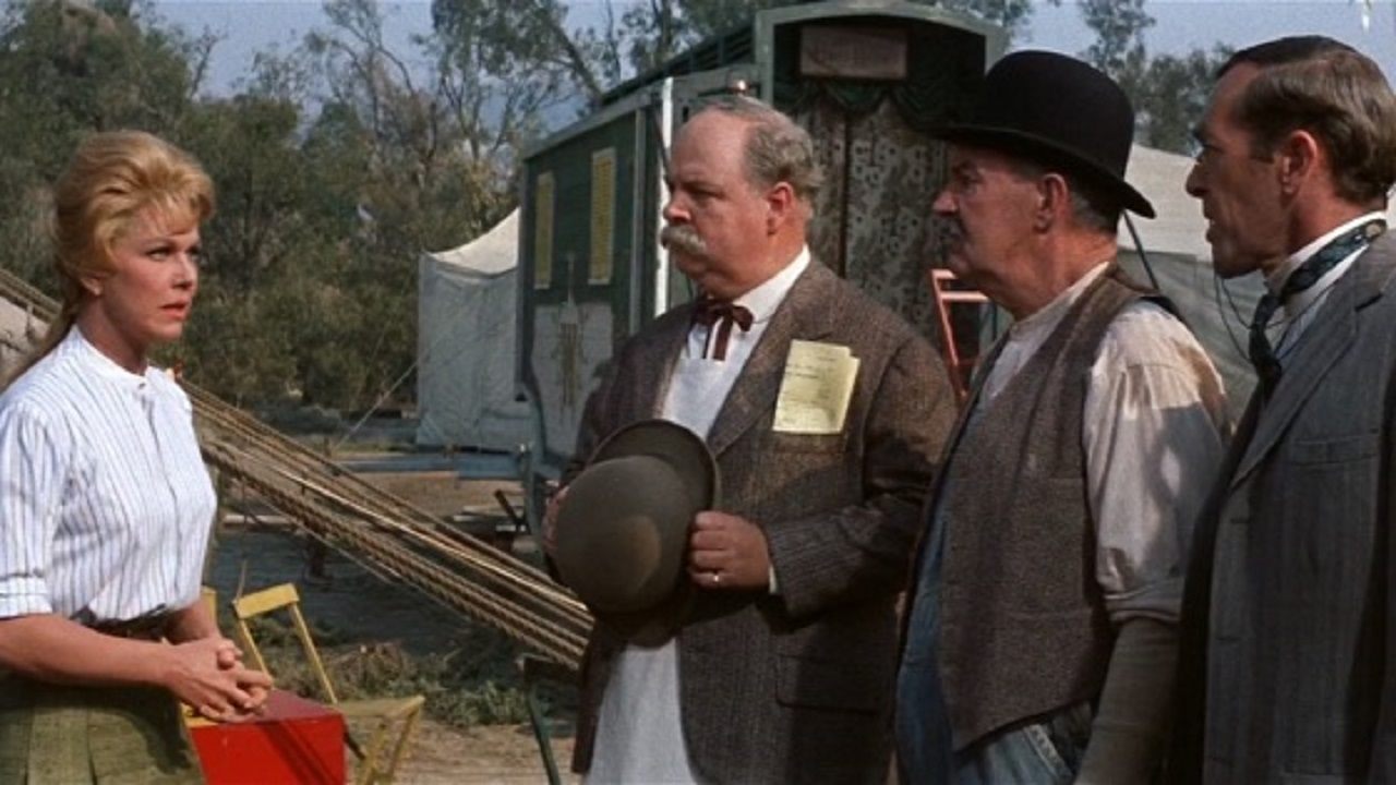 A woman in a striped blouse faces three men dressed in period attire, standing outdoors near tents and trees. One man holds a bowler hat.