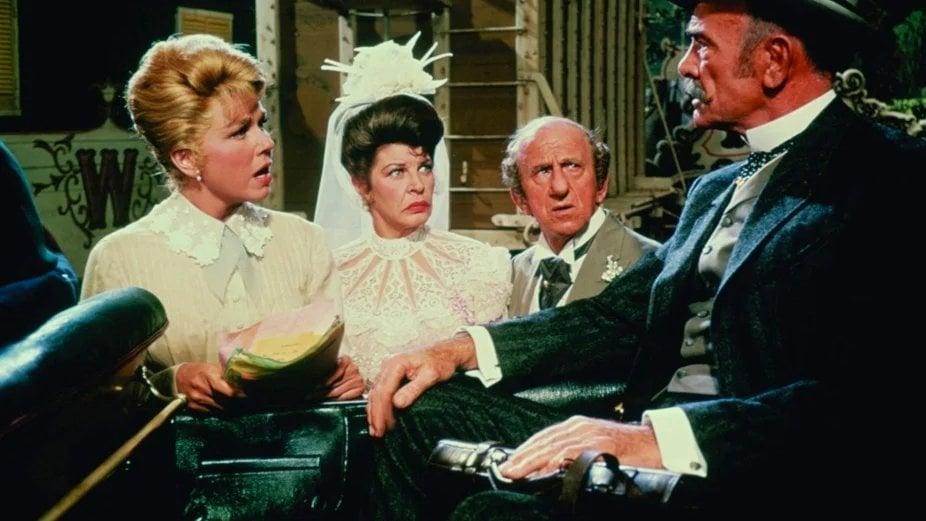 A group of four people wearing period costumes sit together, with one man holding a top hat. The expressions range from serious to surprised.