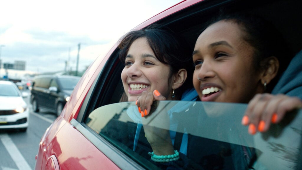 Two people smiling and leaning out of a car window on a busy road, with one holding the window edge.