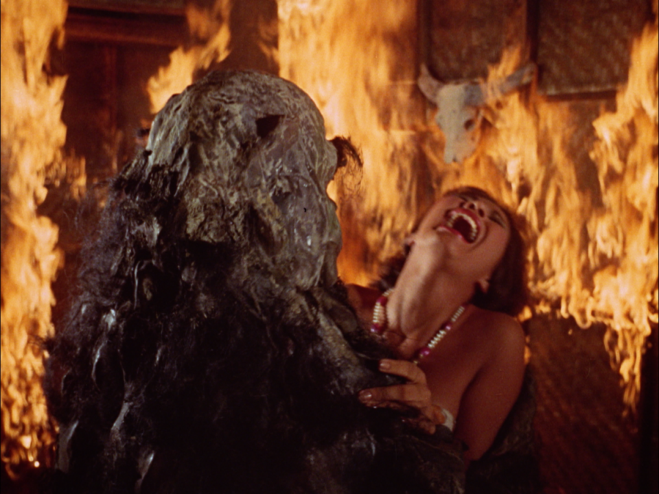 A woman screams while being embraced by a swamp creature inside a room engulfed in flames.