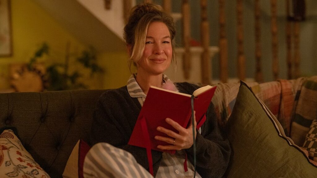 Person sitting on a couch, smiling, and holding an open red book. The setting appears to be a cozy living room.