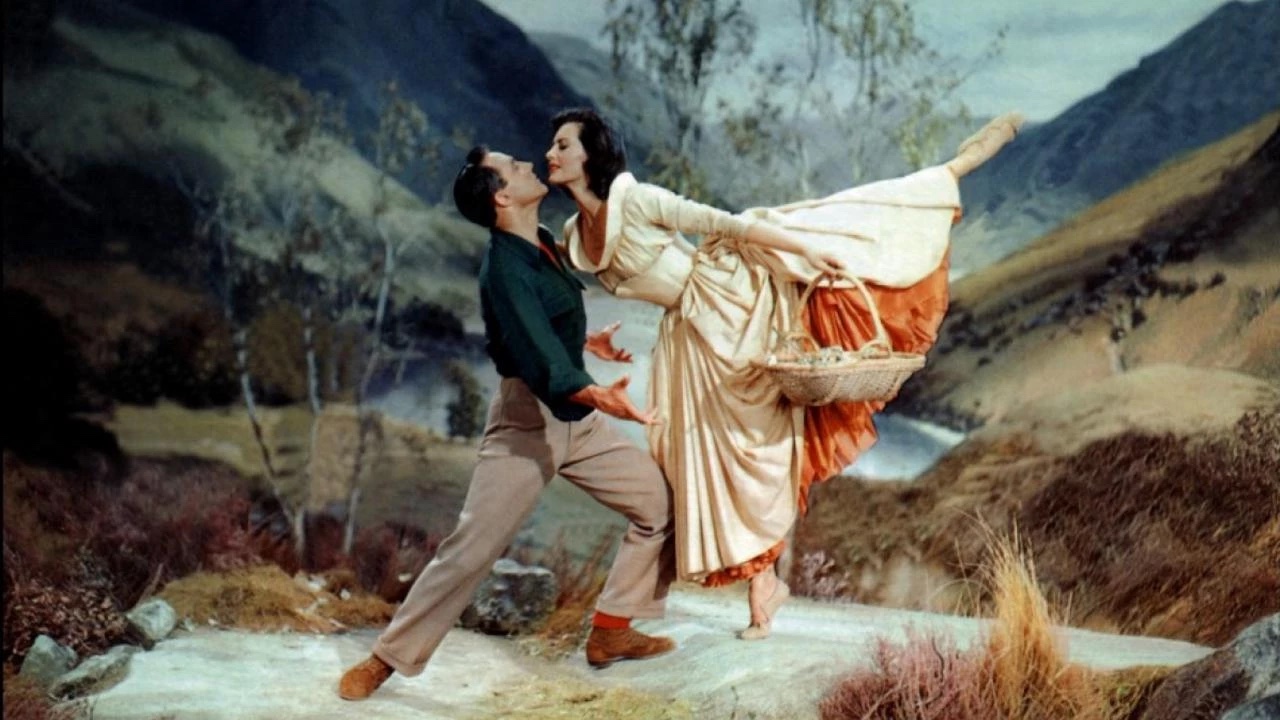 A couple dances outdoors with a mountain landscape in the background. The woman is in mid-air, wearing a long dress, while the man supports her, dressed in casual attire.