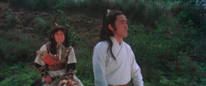 Two individuals in medieval-style attire stand in a grassy outdoor setting, one seated with a hat, the other in a white robe.
