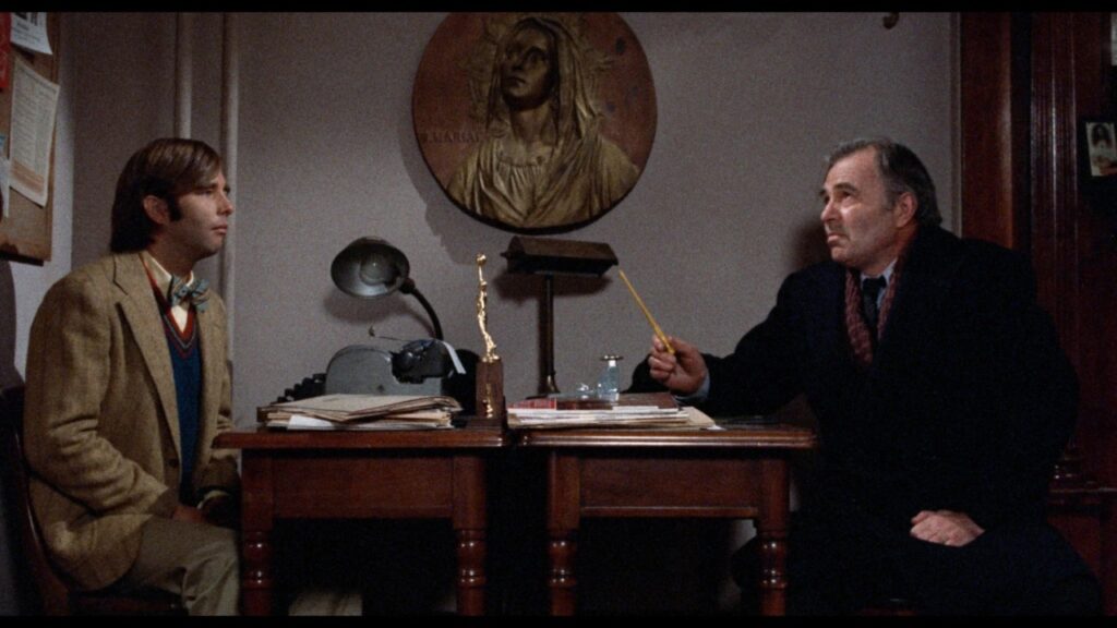 Two men sit across from each other at a desk, in a dimly lit office with a religious image on the wall. One man holds a pencil, and papers are scattered on the desk.