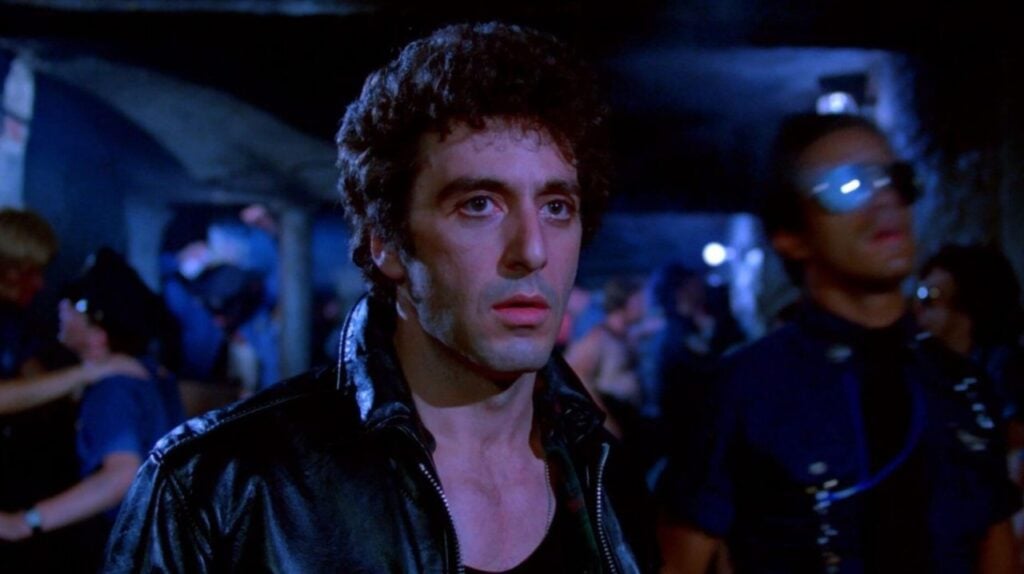 A man with curly hair and a leather jacket stands in a dimly lit room with people behind him.