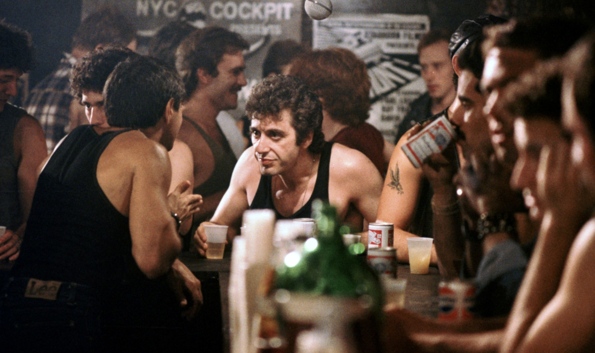 A crowded bar scene with people in tank tops conversing and holding drinks, surrounded by posters and beer cans on the counter.