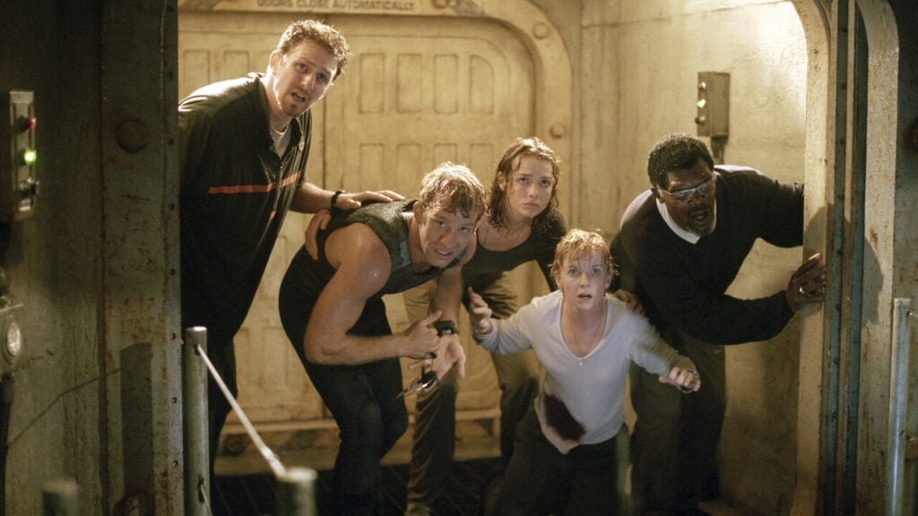 Five people in a dimly lit room, looking intently forward with expressions of urgency. They are standing close together near a doorway.