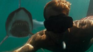 A person wearing goggles takes a selfie underwater while a shark with an open mouth swims nearby.