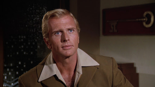 A person with blonde hair and a serious expression is wearing a brown jacket with a wide collar, in a room with a window and a large key decor on the wall.