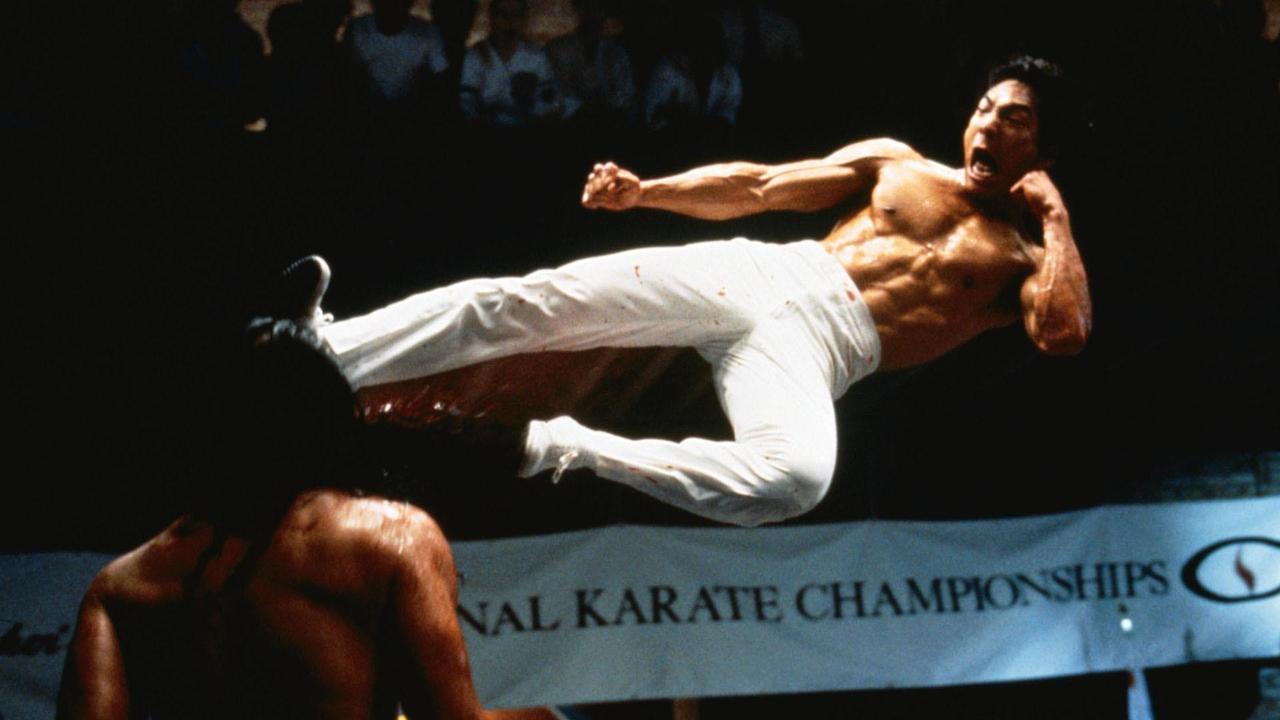 Person performing a high jump kick in a martial arts competition, with another person partially visible below.