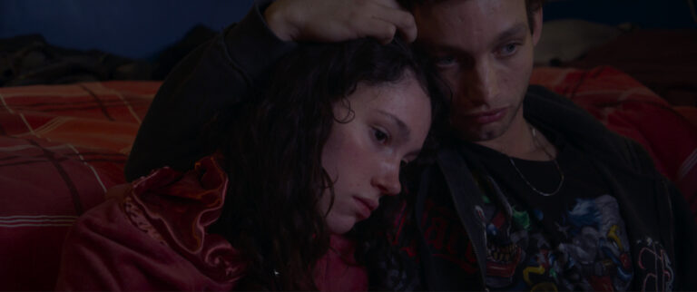 A man and a woman sitting closely on a bed, the man resting his hand on the woman's head. They look contemplative.