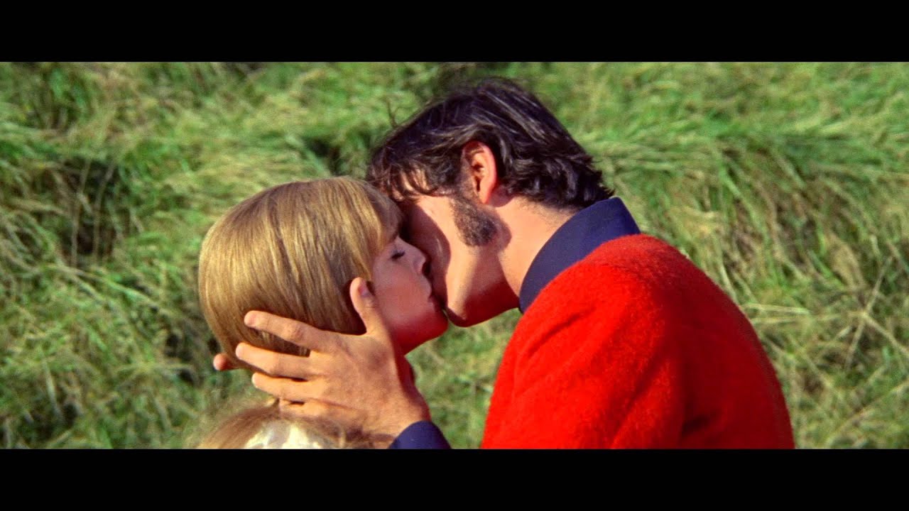 A man and woman kissing outdoors, with the man wearing a red jacket and holding the woman's head gently. Background shows green grass.