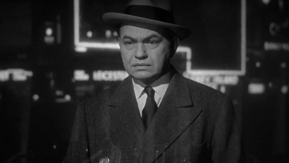 A man in a suit and hat stands in a dimly lit environment, looking serious. The background features blurred, bright lights.