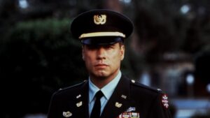 Person in military uniform standing outdoors, wearing a peaked cap, with various decorations on the uniform.