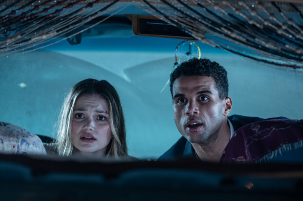 Two people inside a car look surprised, staring forward through the windshield. The interior is dimly lit, with decorative strings hanging above them.