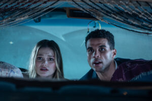 Two people inside a car look surprised, staring forward through the windshield. The interior is dimly lit, with decorative strings hanging above them.