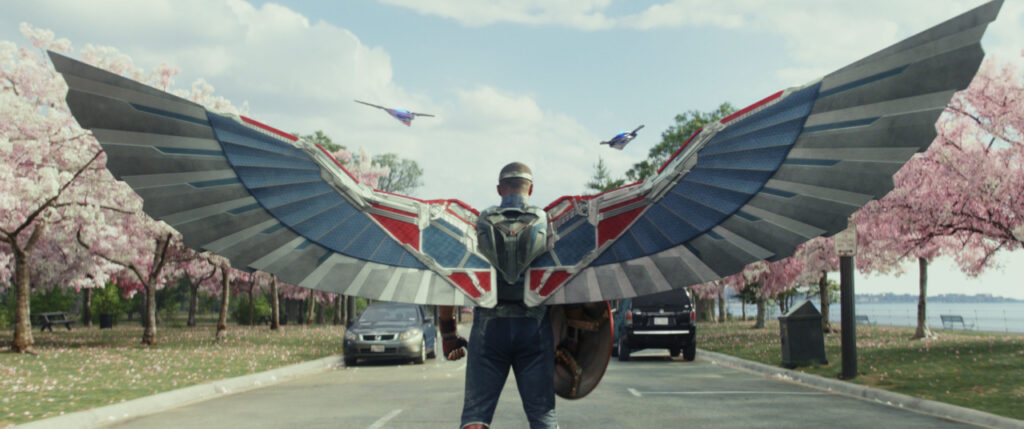 Person with large metal wings stands on a road lined with cherry blossom trees under a clear sky.