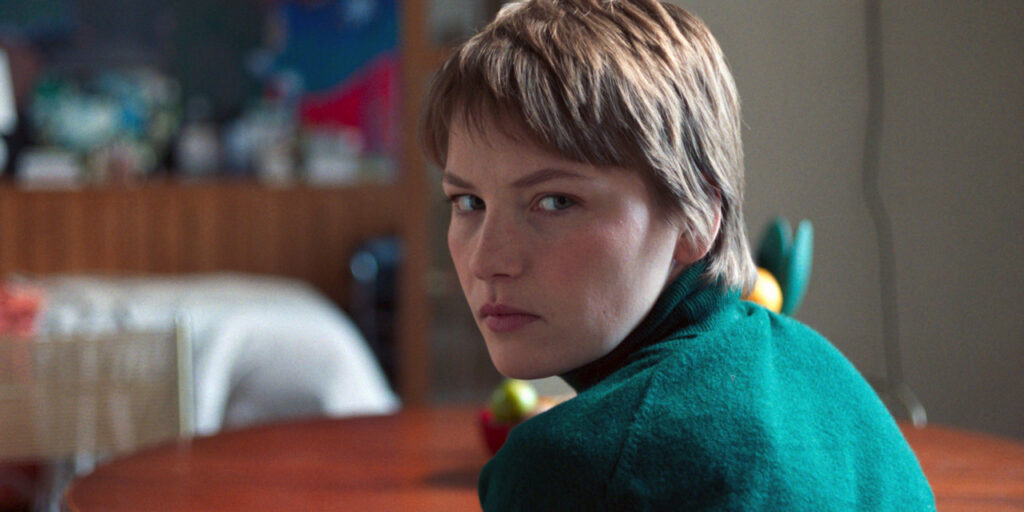 Person with short hair in a green sweater, sitting at a table, looks over their shoulder. Background includes a blurred room with muted colors.