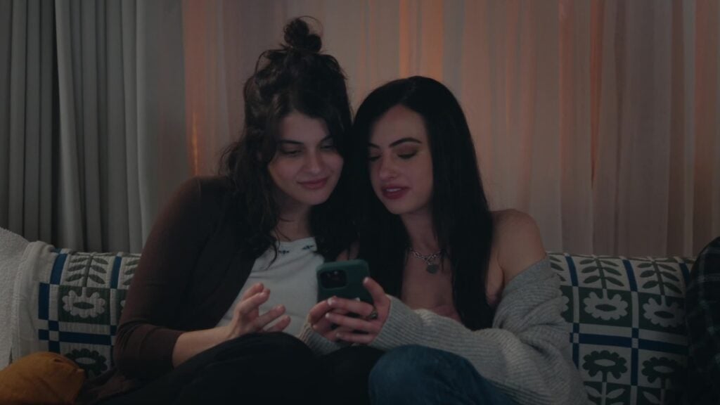 Two people sitting on a couch, looking at a smartphone together. One has curly hair in a bun; the other has straight hair and is wearing a cardigan.