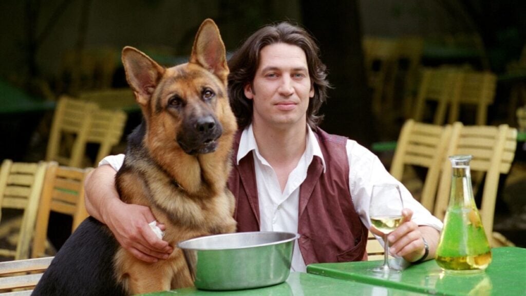 Man sitting at a table with a German Shepherd dog, holding a glass of white wine. A pitcher is also on the table.