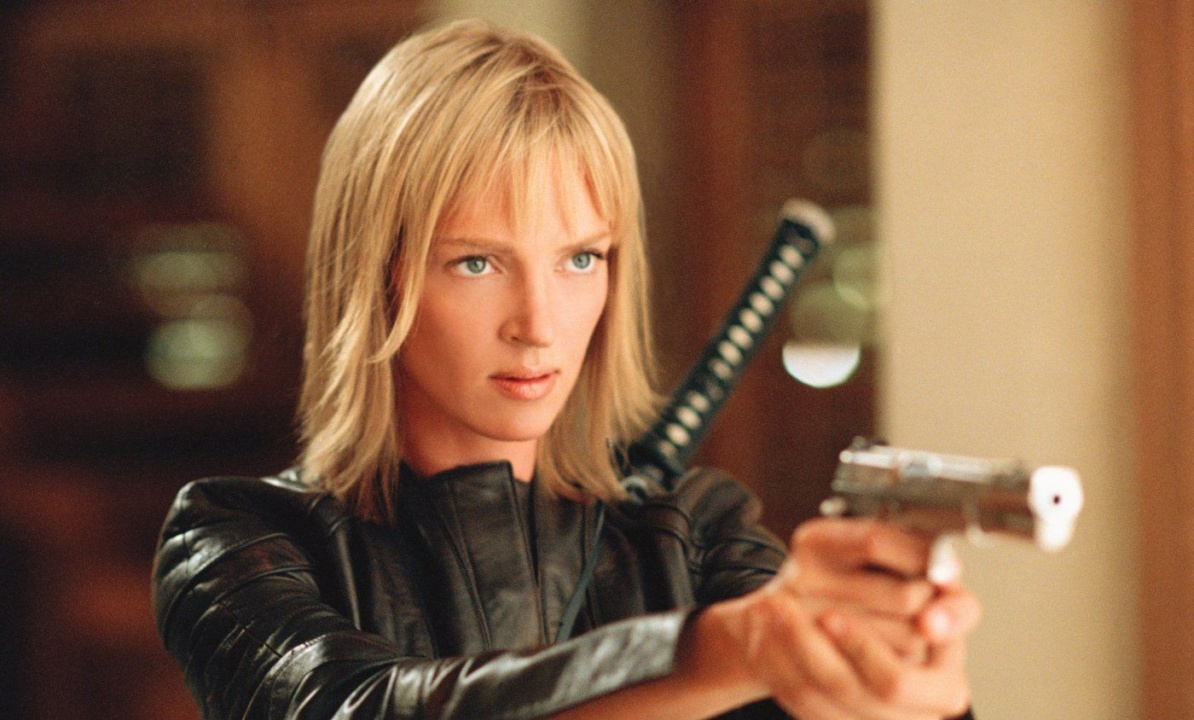 Person with short blonde hair in a black jacket holds a pistol, with a sword on their back, looking intently forward.