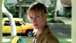 Person with blonde hair and bangs wearing a brown jacket stands outdoors in a suburban area with a yellow car in the background.