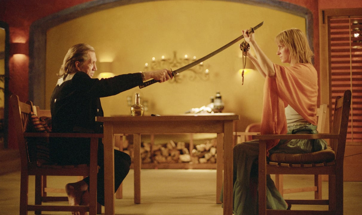 Two people sit at a table in a room, facing each other with swords extended, one wooden and the other metal. The setting includes warm lighting and a decorated background.