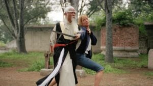 ‘Kill Bill: The Whole Bloody Affair’ 4K UHD Blu-Ray Review – Tarantino’s Fabled Cut Comes Home A person in traditional martial arts attire holds a sword near the neck of another person wearing casual clothes, set outdoors with trees and old stone structures in the background.