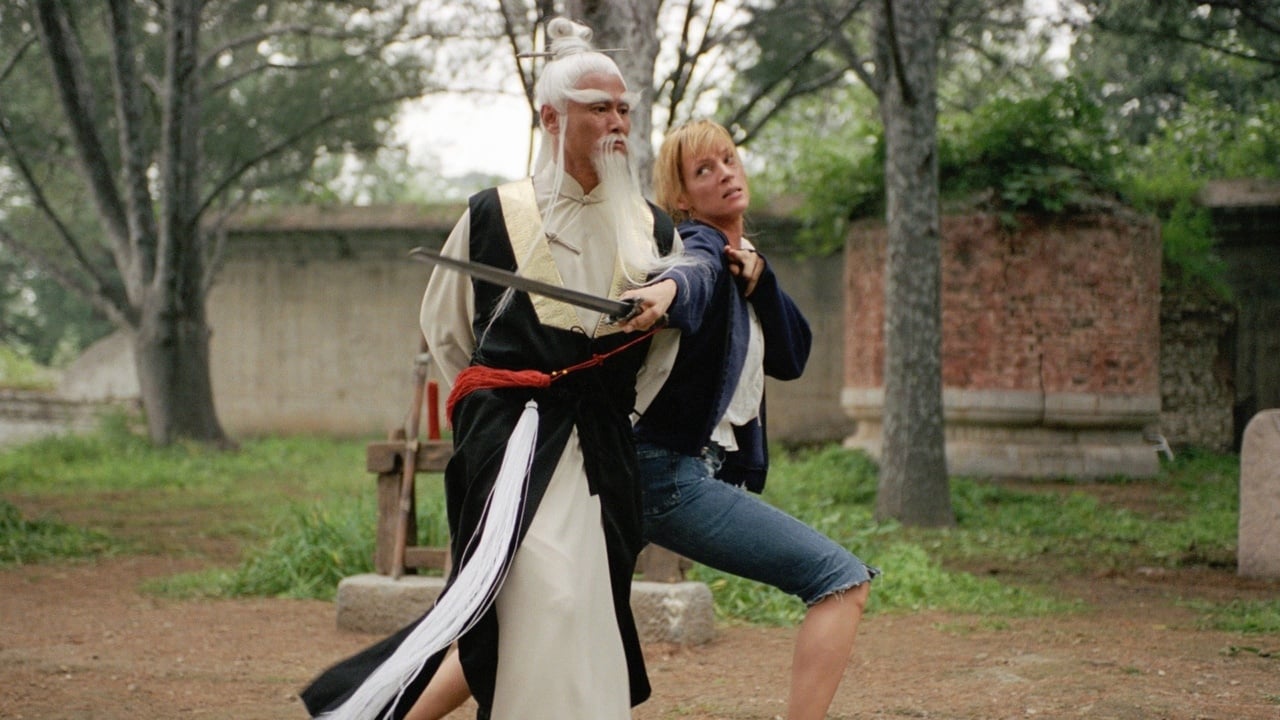 A person in traditional martial arts attire holds a sword near the neck of another person wearing casual clothes, set outdoors with trees and old stone structures in the background.