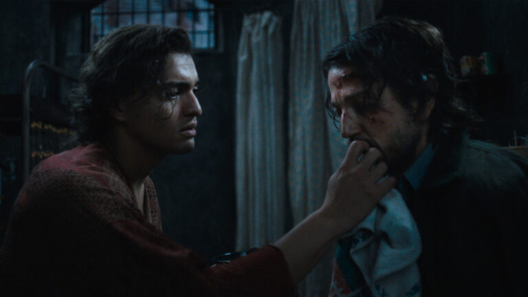 Two men in a dimly lit room, one tending to the other's facial injuries with a cloth, appearing serious and concerned.