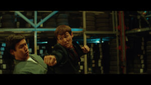 Two men are engaged in a fight in a warehouse stocked with tires. One man is throwing a punch while the other is reacting to it.
