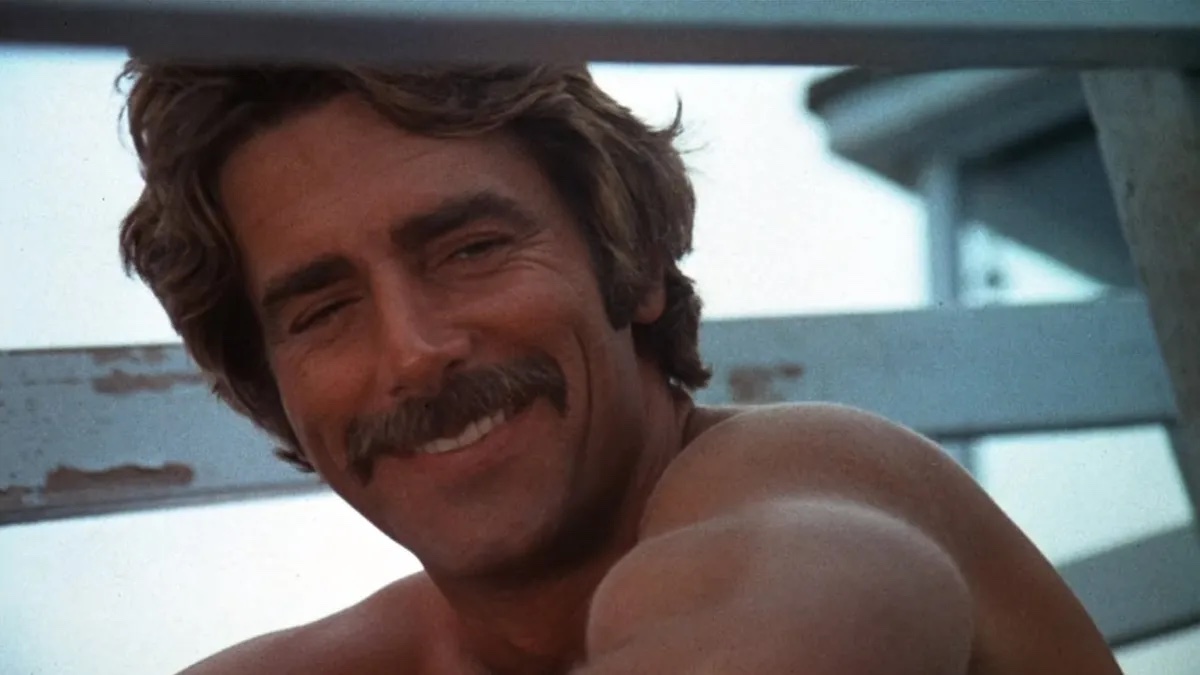 A person with a mustache and wavy hair smiles while sitting shirtless outdoors.