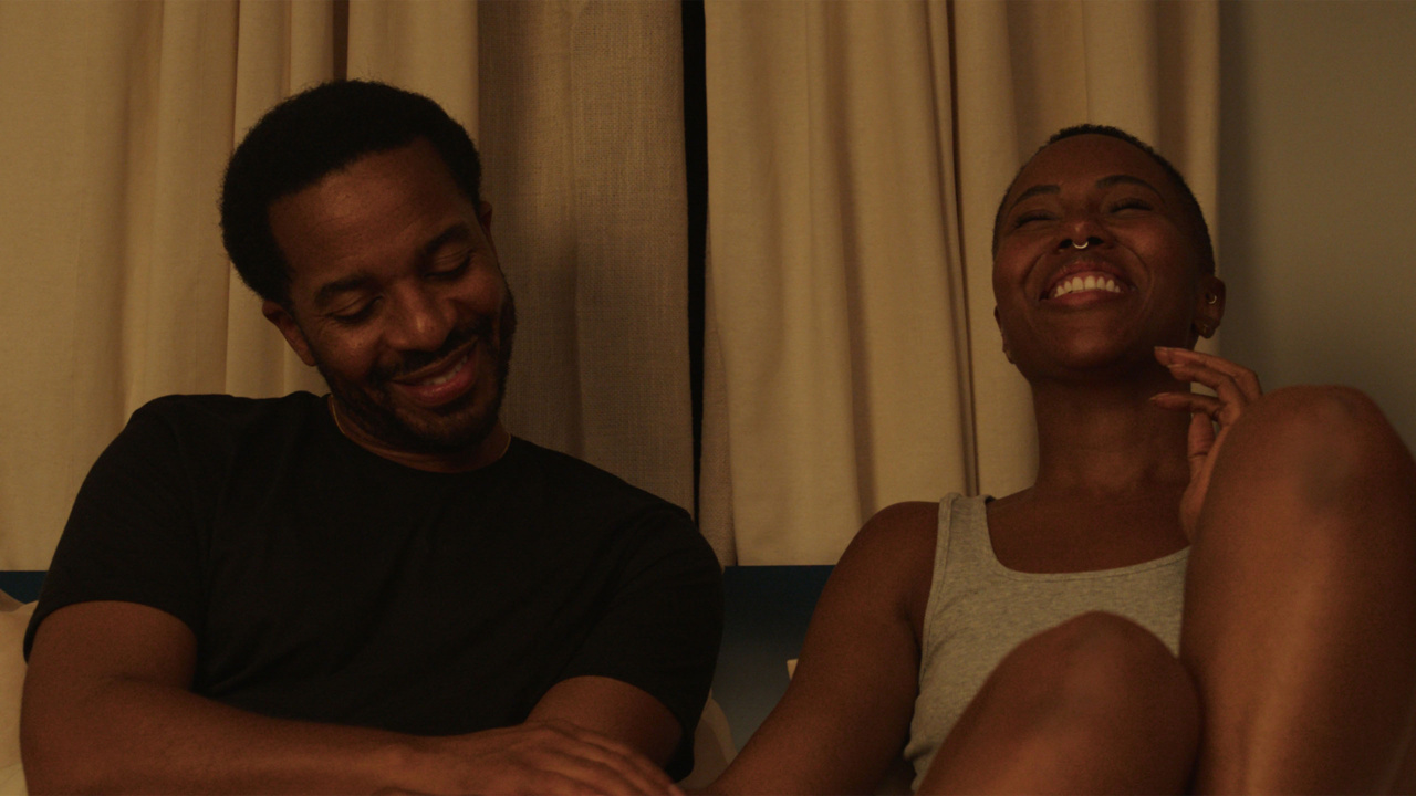 A man and a woman sitting together on a couch, smiling and sharing a light moment. The room has beige curtains in the background.