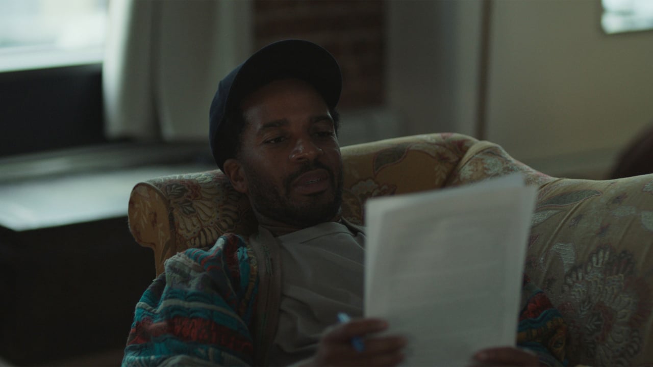 Person wearing a hat and colorful sweater, sitting on a patterned sofa, reading a document with focused expression.