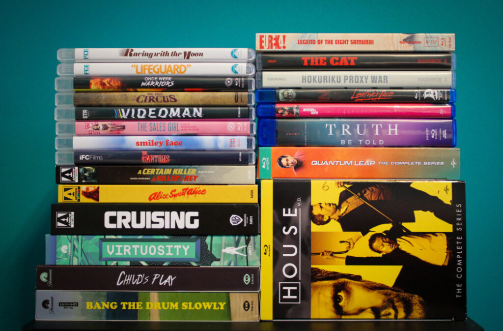 A collection of DVD and Blu-ray cases stacked against a teal background, featuring various movies and TV series.