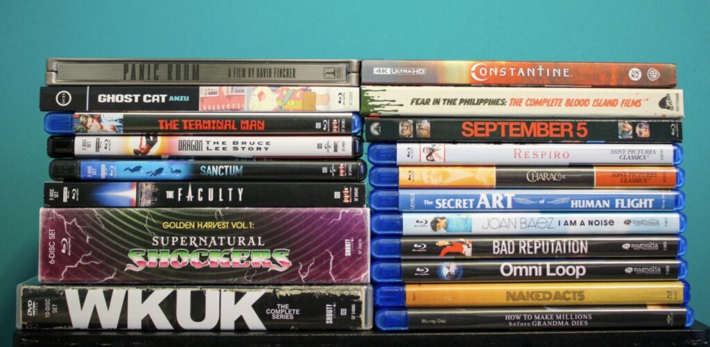 Stacks of DVD and Blu-ray cases against a teal background, featuring various titles including "Constantine," "Ghost Cat," "The Terminal Man," and "Supernatural Shockers.