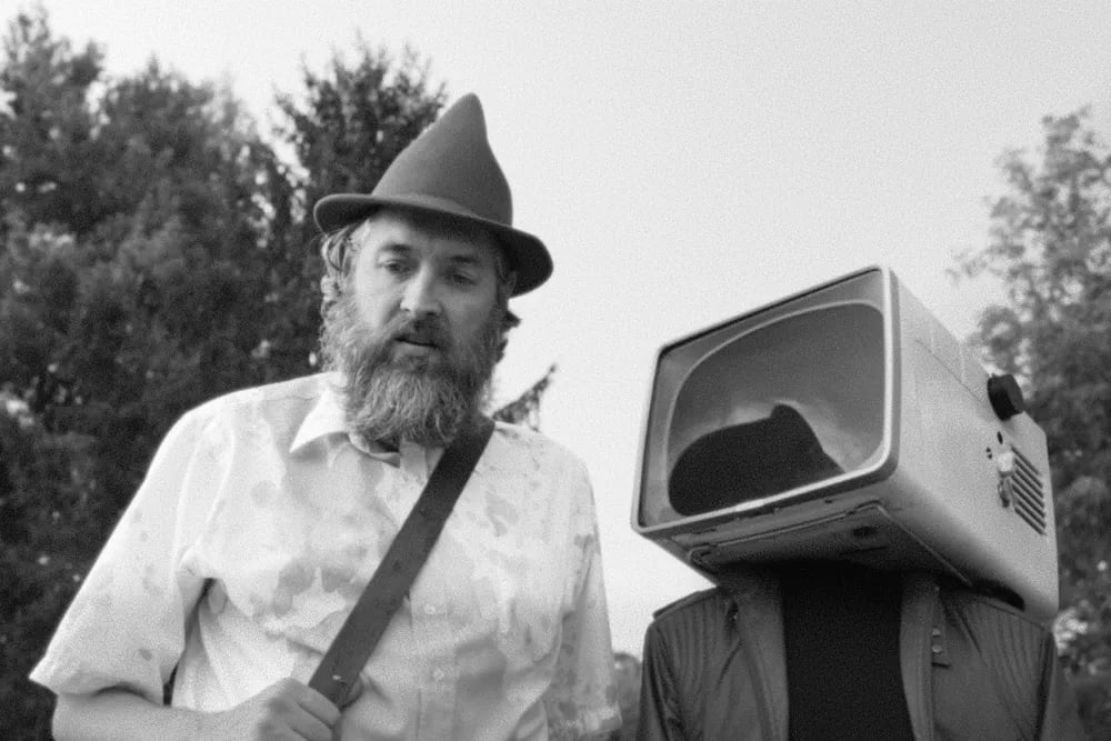 Person with beard in a hat next to another person with an old TV on their head, outdoors.