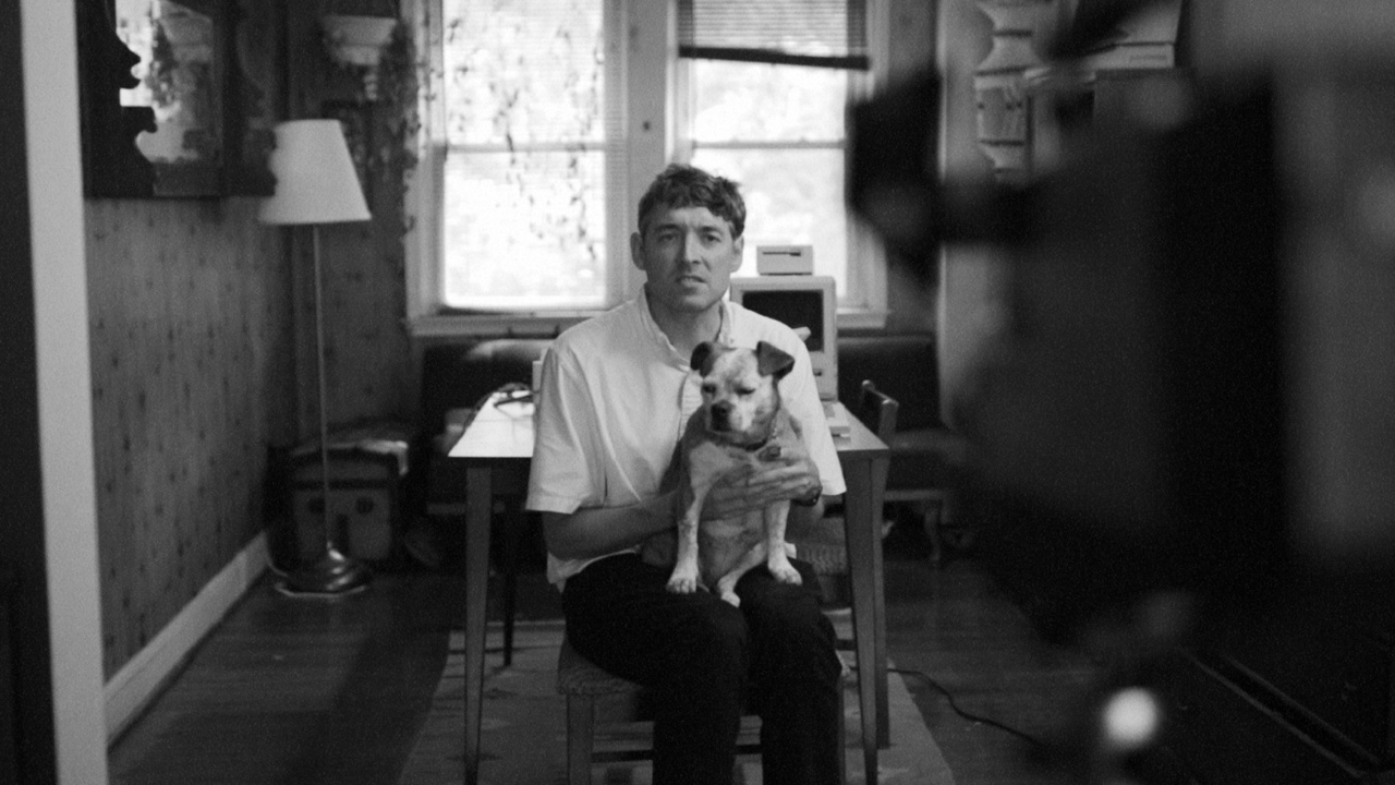 Person sitting on a chair indoors, holding a small dog. The room has a lamp and a desk by the window. Image captured in black and white.