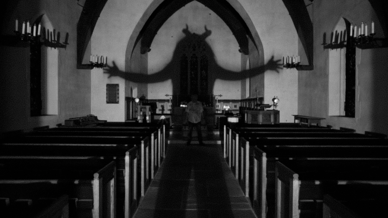A person stands in a dimly lit church casting a large, menacing shadow with outstretched arms that resembles a creature with horns.