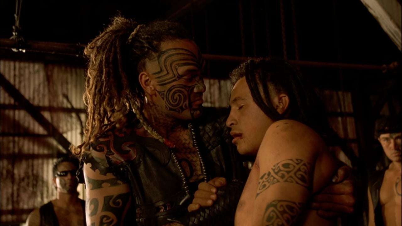 Two people with tribal tattoos face each other closely in a dimly lit setting. One person is holding the other by the shoulder. Others with similar tattoos stand in the background.