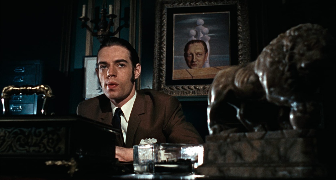 A man in a suit sits at a desk with a horse statue, looking at the camera. A painting with a head and spheres is on the wall behind him.