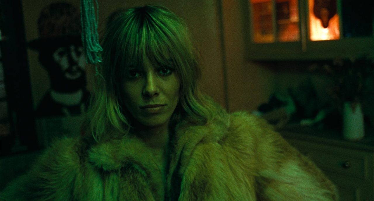 Person with blonde hair in a fur coat stands in a dimly lit room with green lighting. A painting and a vase are visible in the background.