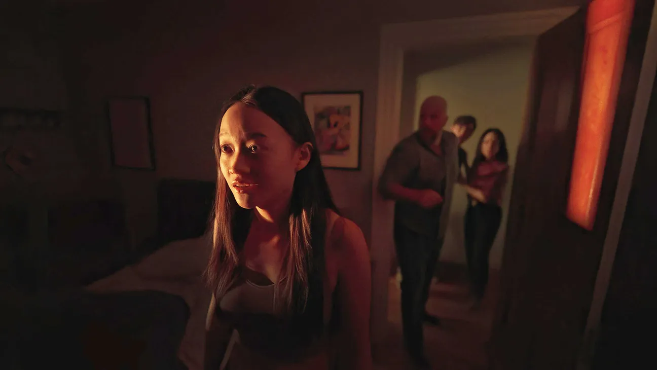 A woman stands in a dimly lit room, illuminated by warm light. Three people are positioned in the doorway behind her.