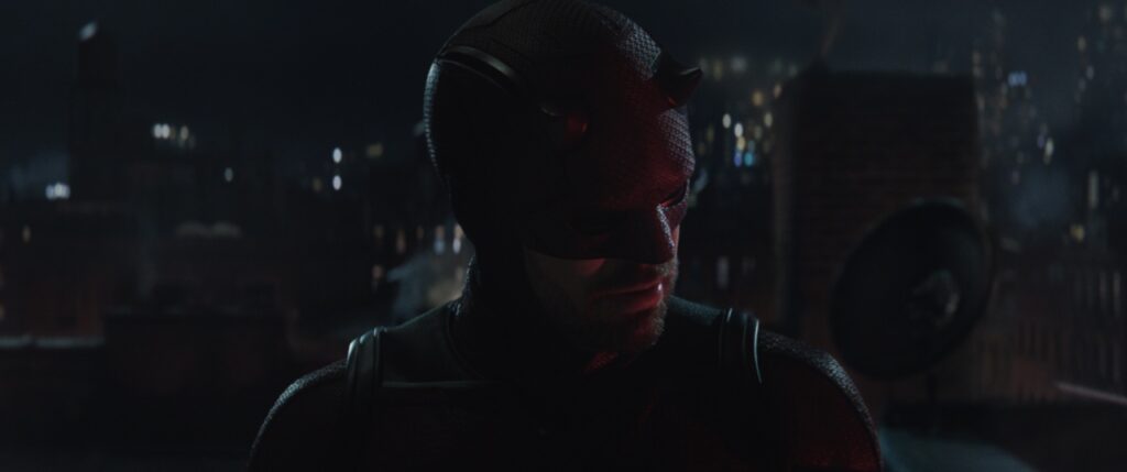 A masked person in a dark costume stands on a rooftop at night with a city skyline in the background.
