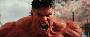 A red-skinned, muscular figure with short dark hair appears to be shouting in an outdoor setting with blurred trees in the background.